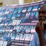 Fisherman Amaladasan stands beside a poster showing photos of 70 fishermen missing from Vallavilai village in Kanyakumari district since Cyclone Ockhi. (Photo by M M Paniyil)