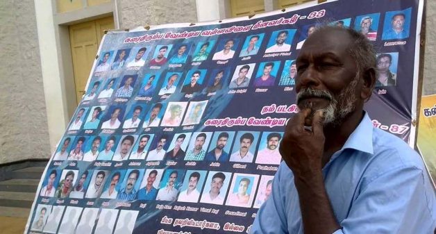 Fisherman Amaladasan stands beside a poster showing photos of 70 fishermen missing from Vallavilai village in Kanyakumari district since Cyclone Ockhi. (Photo by M M Paniyil)